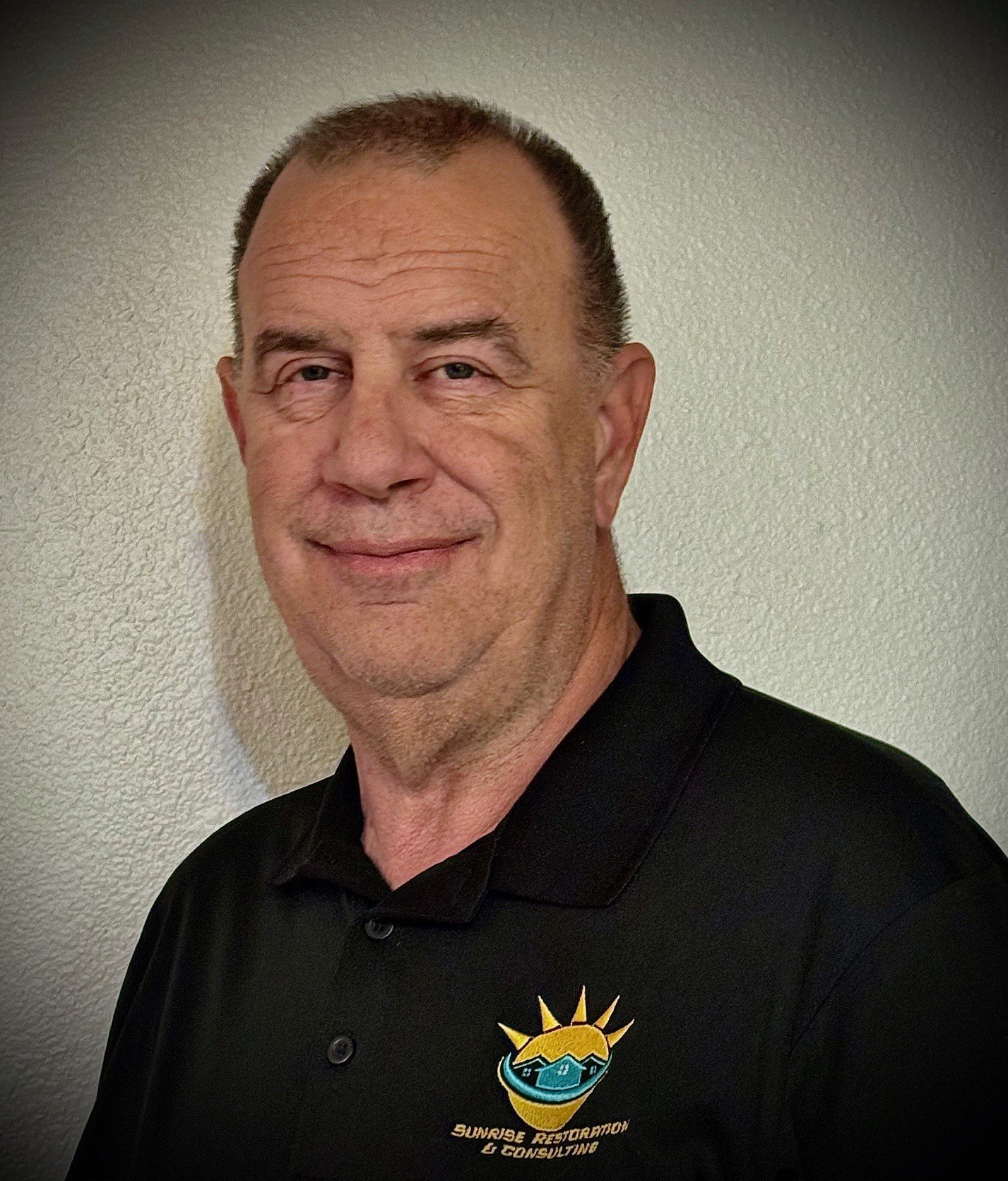 Man wearing a black polo shirt with "Sunrise Restoration & Consulting" logo, standing against a light-colored wall.