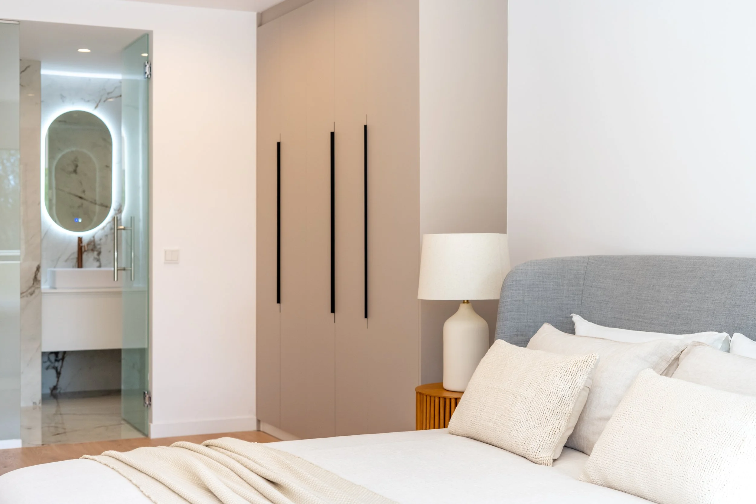 A bedroom with a bed, beige pillows, a beige blanket, a wooden nightstand with a white lamp, a tall beige wardrobe with black handles, and a view into a bathroom with a marble sink and a backlit mirror.