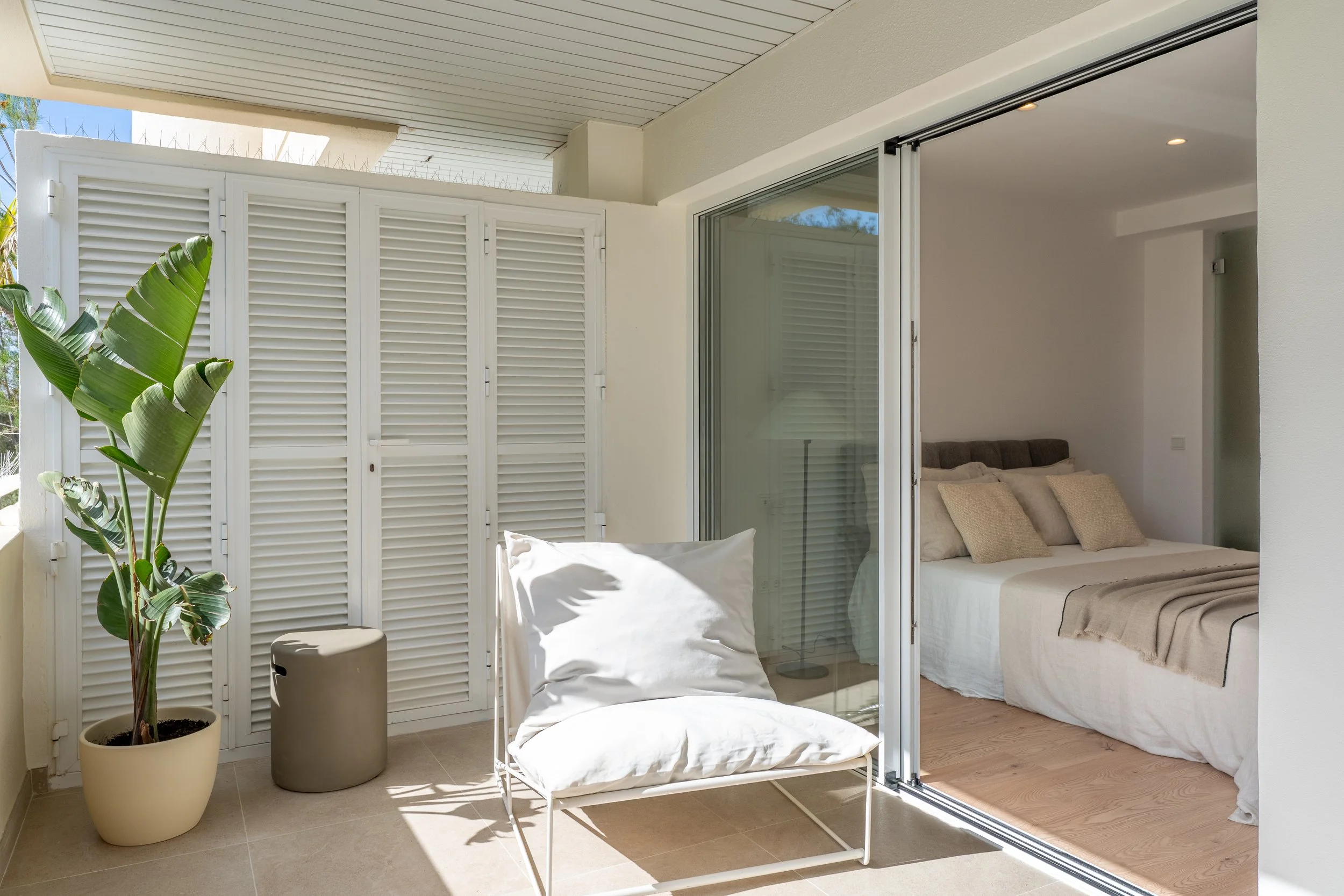 Balcony with a potted plant, a white lounge chair with a pillow, and a sliding glass door leading into a bedroom with a bed and beige pillows.