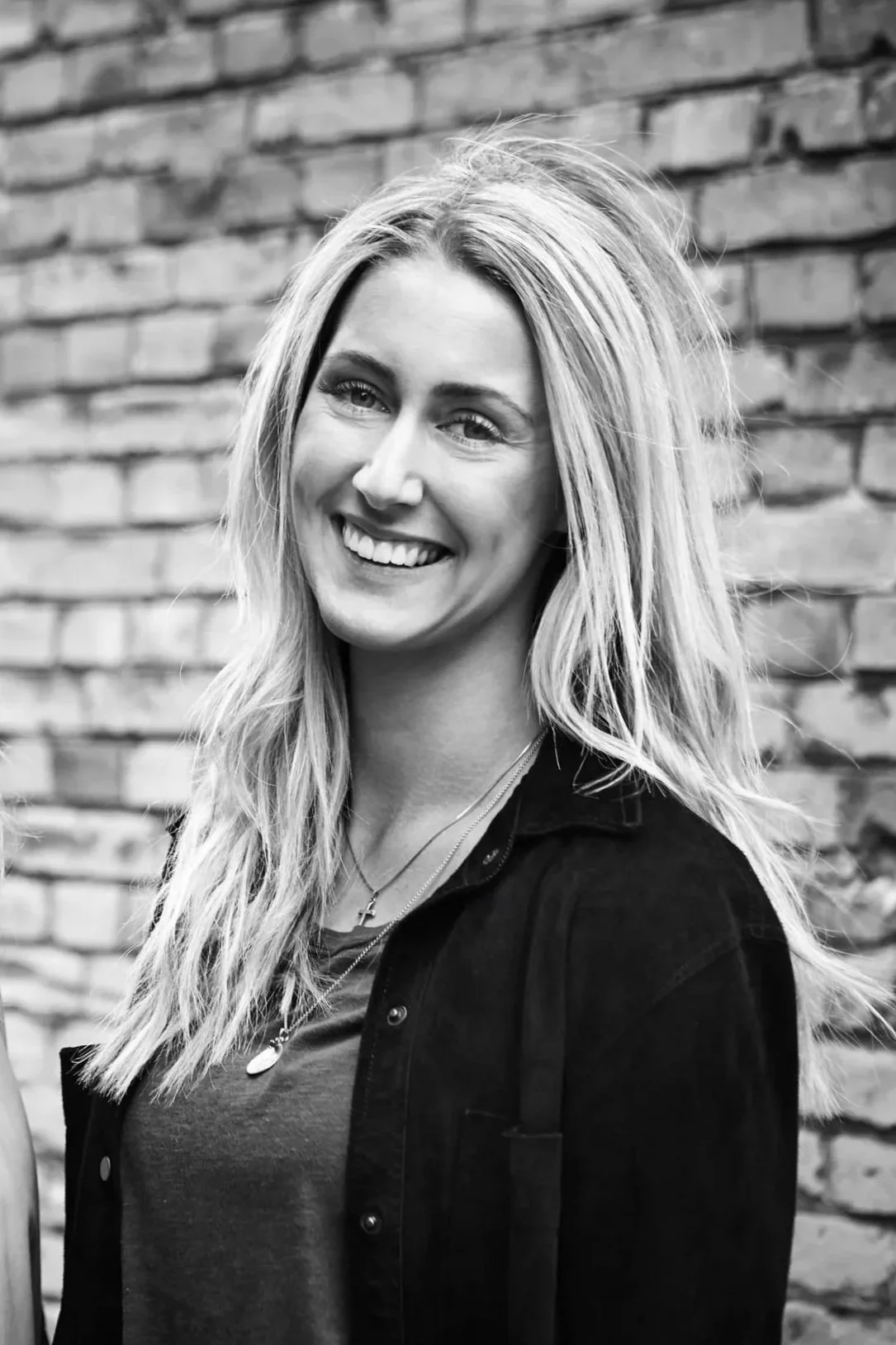 Sort-hvid portræt af smilende blond kvinde med festligt hår, smaragdfarvet t-shirt, sort jakke og kors halskæde foran en murstensvæg.