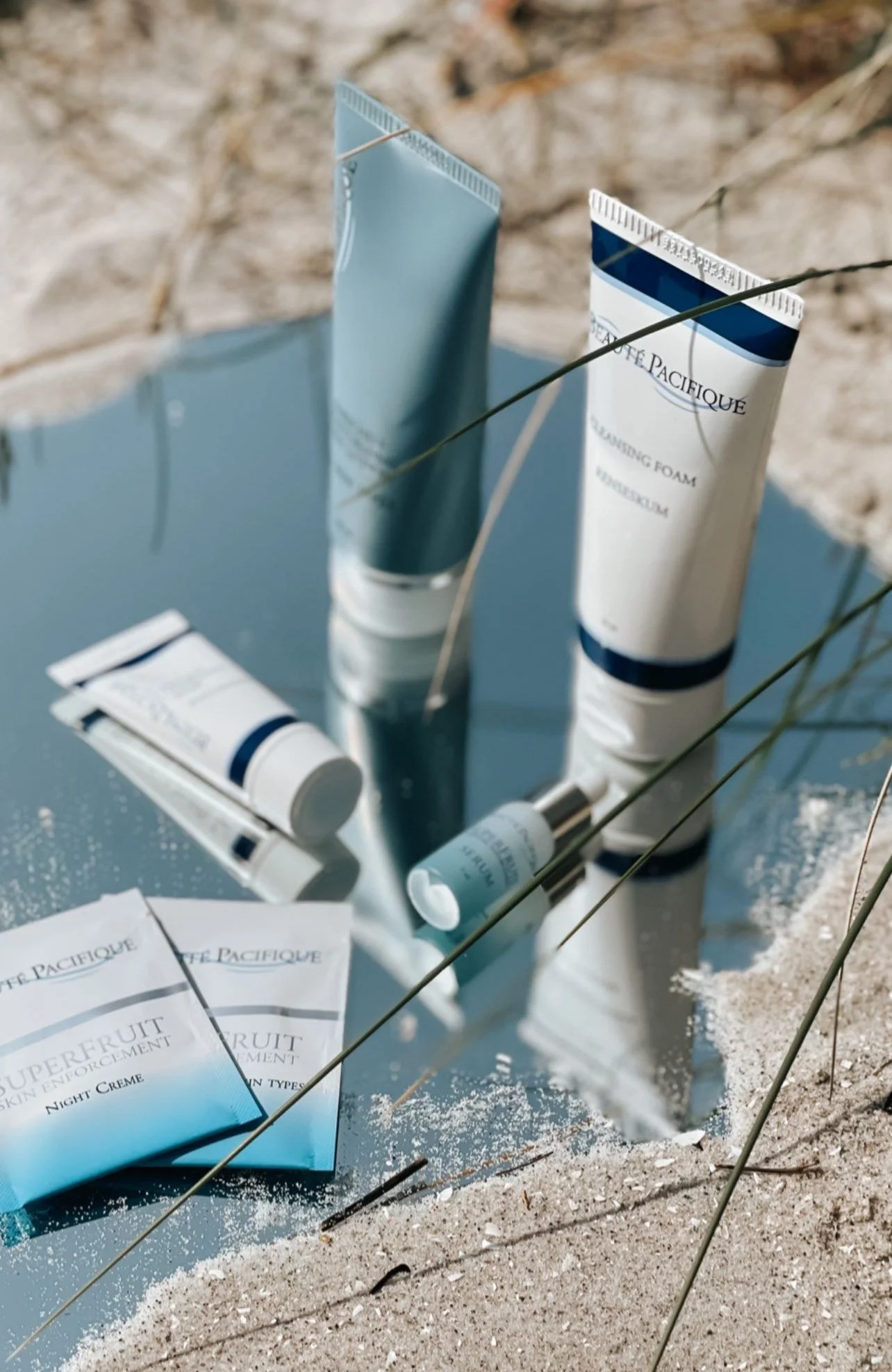 Skønhedsprodukter som ansigtscreme, rensningsskum og serum på en spejlflade omgivet af sand og tørt græs.