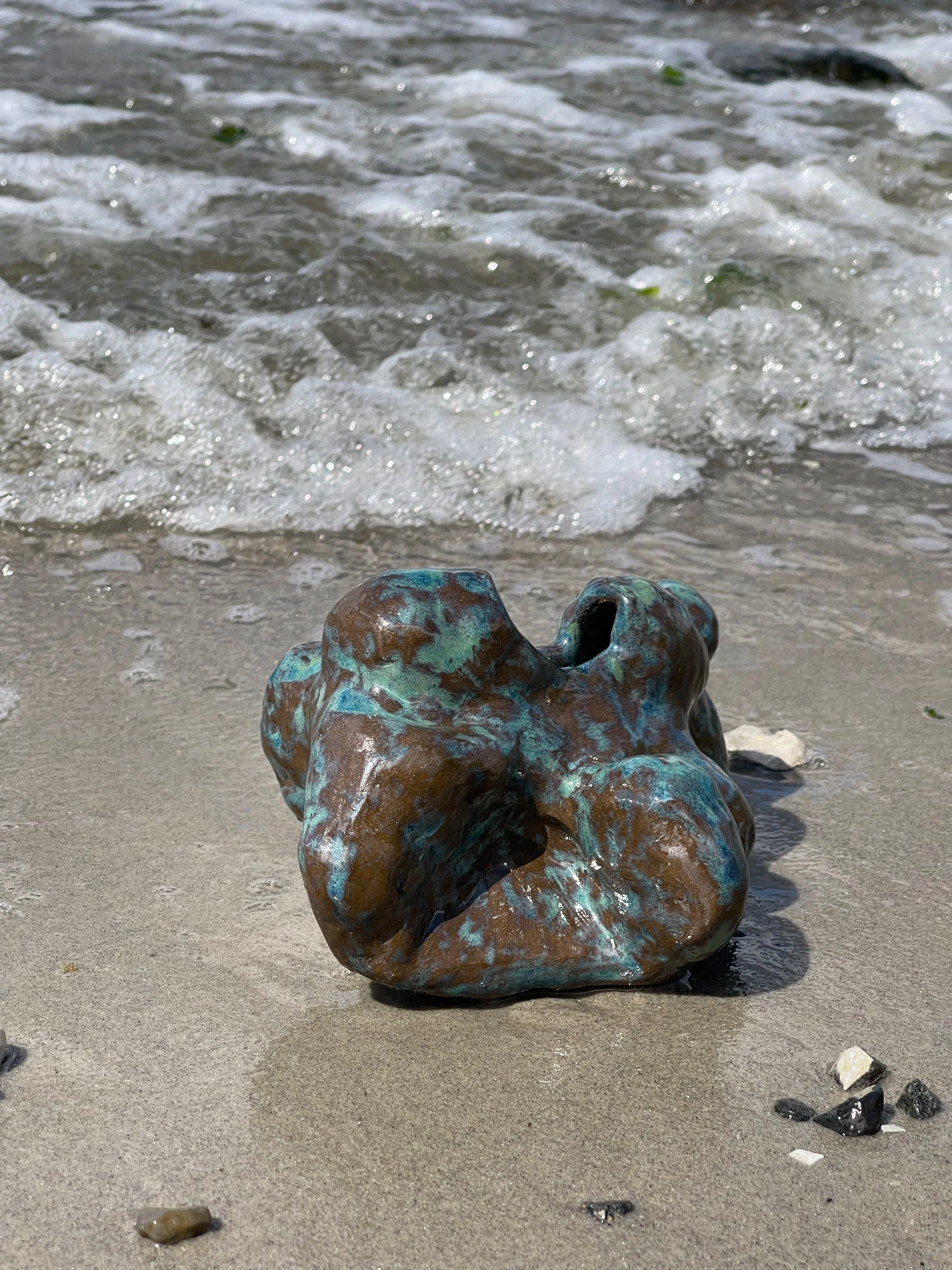 Metal skulptur formet som en abstrakt, organisk form på sandstrand med havbølge i baggrunden.
