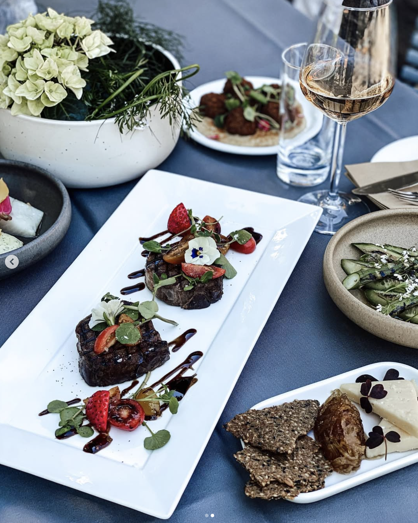 En tallerken med tre stykker kød med flødestuvede grøntsager, garneret med frugt, urter og balsamico reduction. Rundt omkring er der forskellige små fade med salater, brød, og drikkevarer.