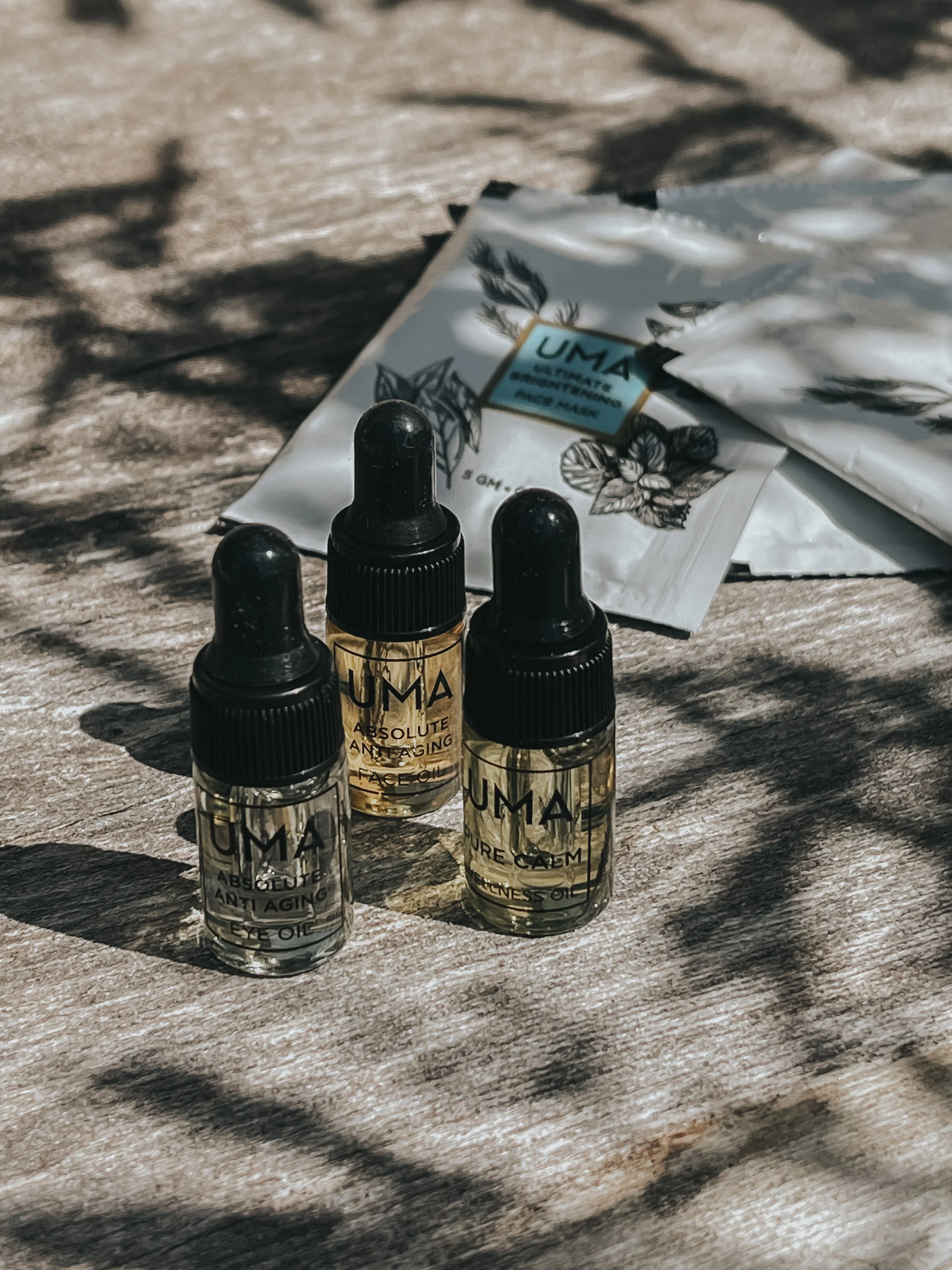 Tre små flasker med æteriske olier og serienummer, samt nogle ampuller i poser, placeret på en træoverflade med skygger.