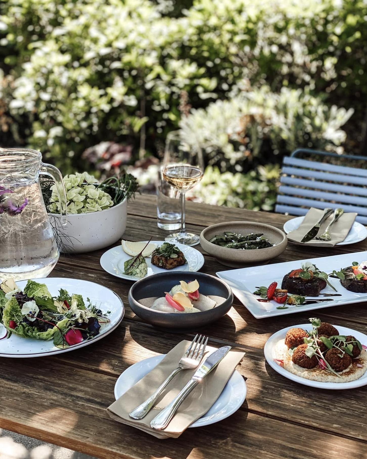 Udendørs restaurantbord med forretter, salat, drikkevarer og bestik, omgivet af grønne planter.