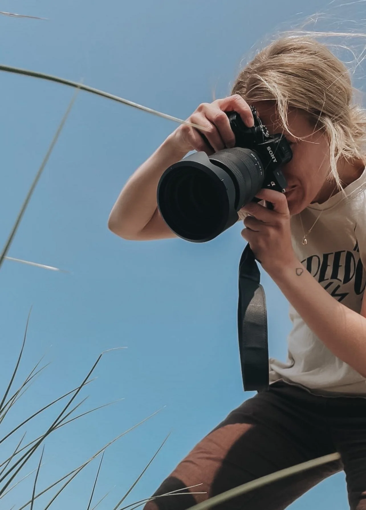 Person med blond hår fotograferer med kamera i udendørs omgivelser under blå himmel.