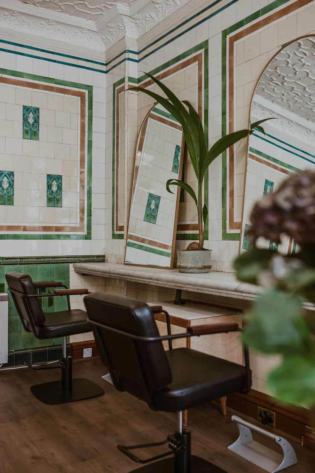 Interior of a room with ornate tiled walls, a large potted plant, two black chairs with wooden armrests, and a mirror leaning against the wall.