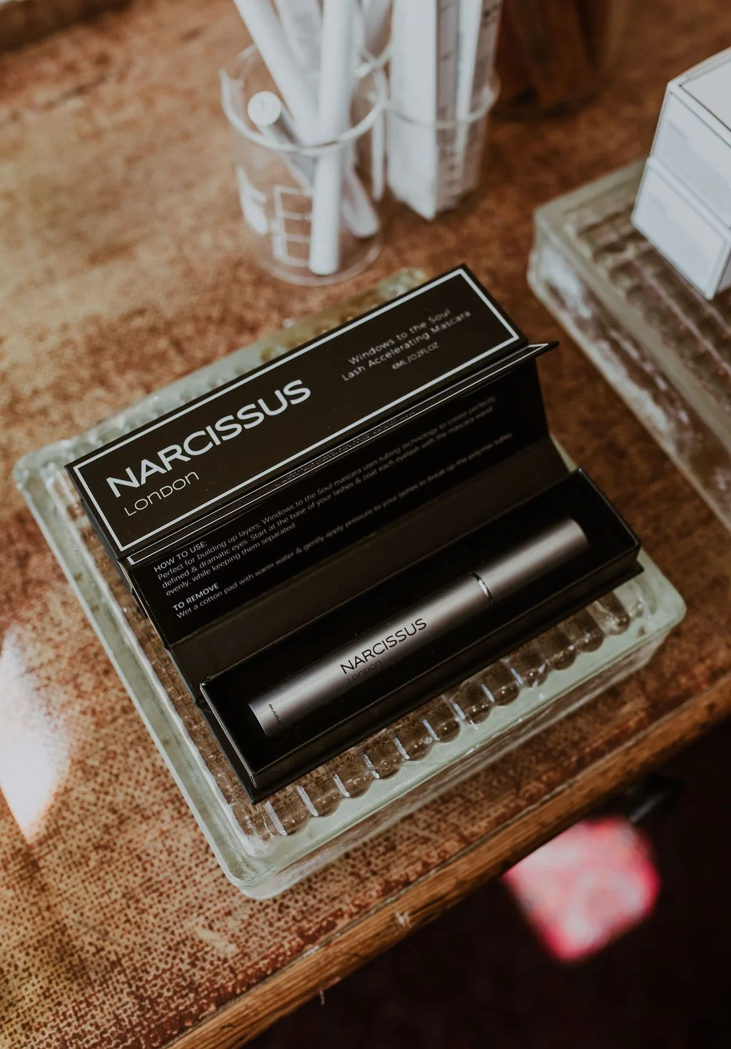 A Narcissus London beauty serum in a sleek silver bottle inside a black box on a glass tray, surrounded by office supplies on a wooden surface.