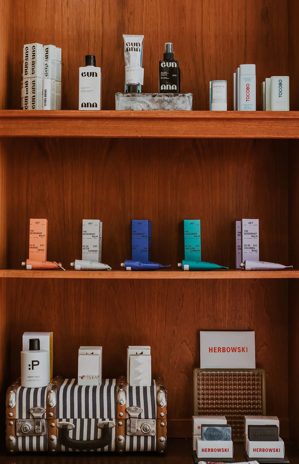 Wooden shelf displaying various skincare or cosmetic products. Top shelf has creams and sprays in sleek white, black, and gray packaging. Middle shelf features small tubes of deodorant balm in different colors, labeled with scent names. Bottom shelf shows a striped cosmetic bag, a white bottle, and a sign with the brand name Herbowksi. Additional small boxes and products are on a table below.