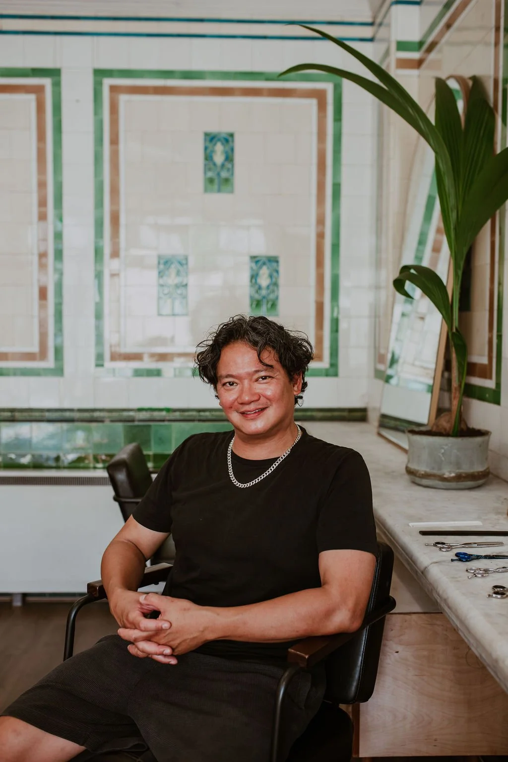 A smiling man with curly dark hair, wearing a black shirt, silver chain necklace, sitting in a black chair in a room with white, green, and brown tile walls, a marble countertop with scissors and tools, and a potted plant nearby.