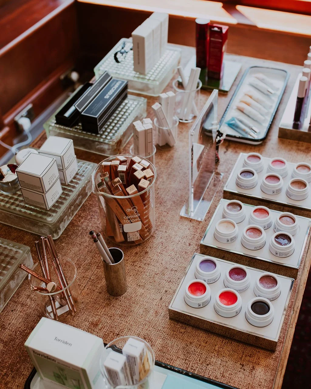 A makeup or skincare product display with various small jars and boxes, brushes, and cosmetic palettes arranged on a wooden table.