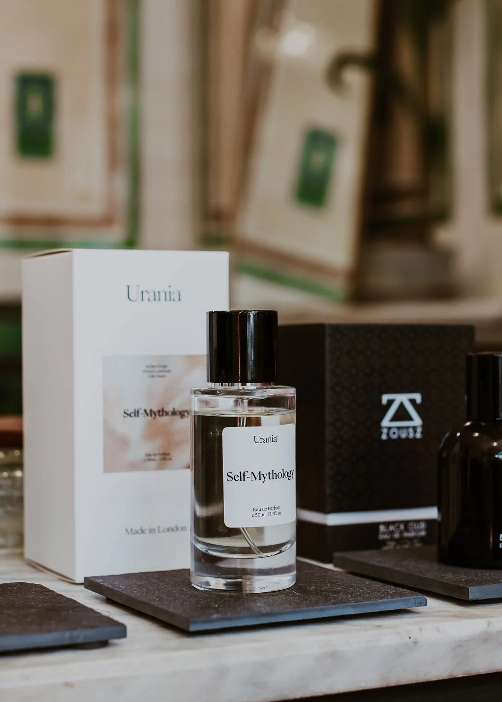 A bottle of Urania Self-Mythology perfume displayed on a black mat with its packaging behind it on a store shelf.