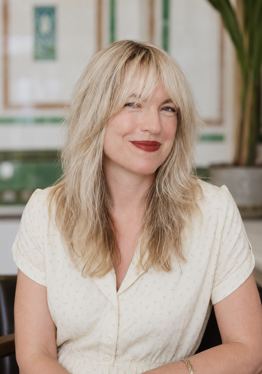 A woman with blonde hair, wearing red lipstick, and a cream-colored blouse with small yellow patterns, smiling at the camera in an indoor setting with a plant and blurred background.