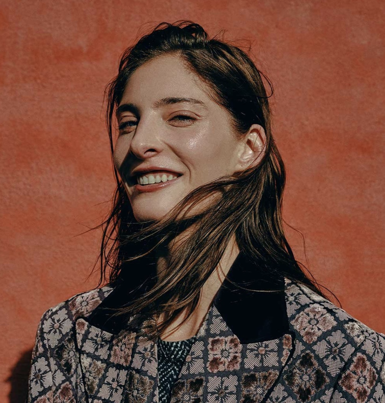A woman with long brown hair smiling against a red textured background, wearing a patterned jacket with floral designs.