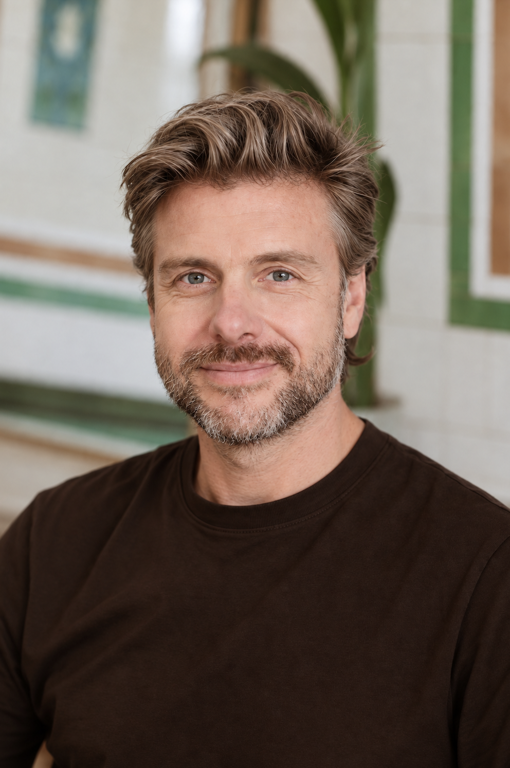 A man with light brown hair, blue eyes, and a beard smiling at the camera, wearing a dark brown shirt, with a green plant and artwork on the wall behind him.