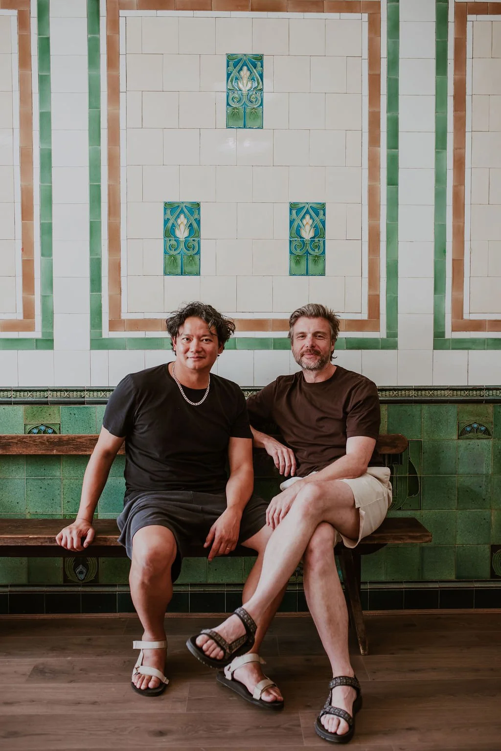 Two men sitting on a wooden bench in front of a decorative tiled wall, one with black hair and a chain necklace, the other with gray hair and a beard, both casually dressed.