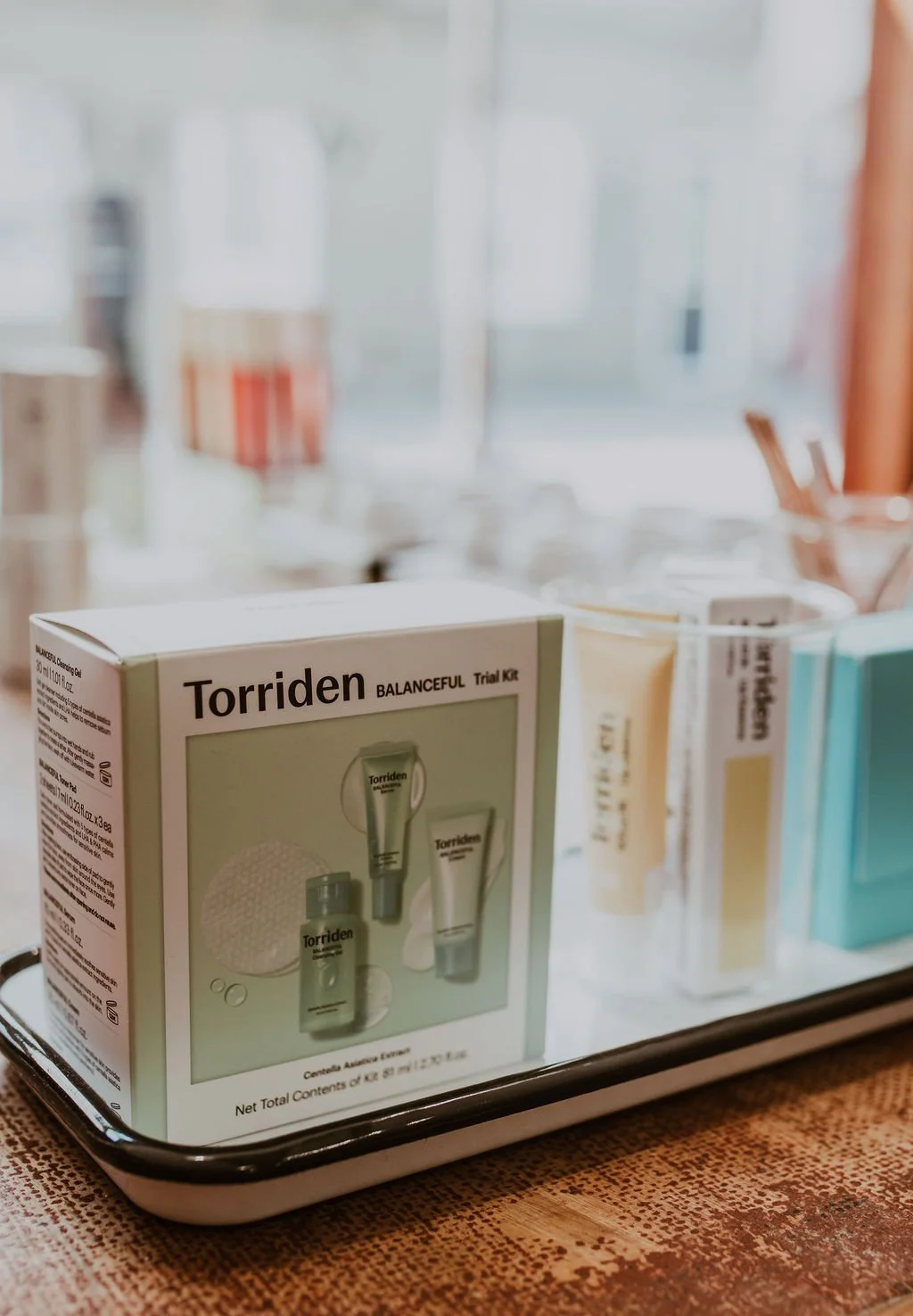 A display tray with a box of Torriden Balanceful skincare trial kit and several other skincare products in bottles and tubes, set on a wooden surface in a bright, blurred background.