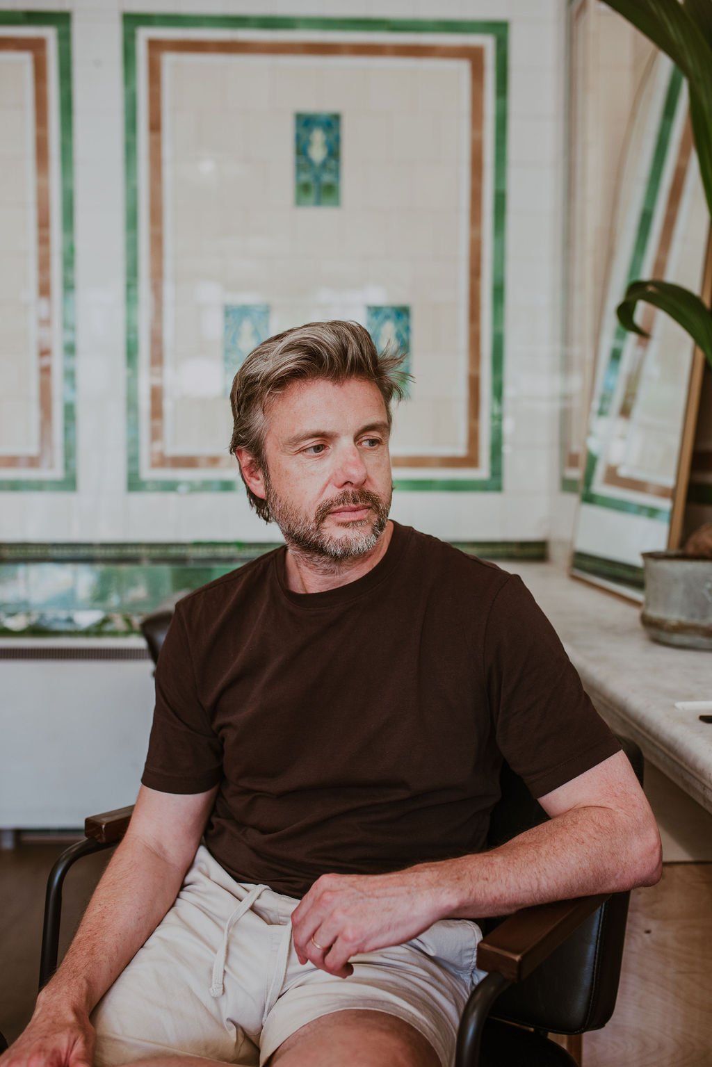A middle-aged man with gray hair and beard, wearing a black t-shirt and beige shorts, sitting in a chair in what appears to be an art or craft studio with painters' palettes and colorful artwork in the background.