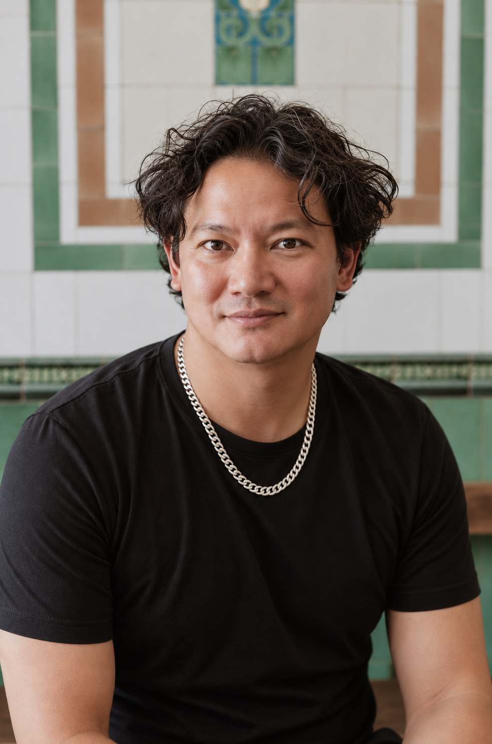 A young man with dark curly hair and a silver chain necklace, wearing a black t-shirt, sitting indoors in front of a decorative tiled wall.
