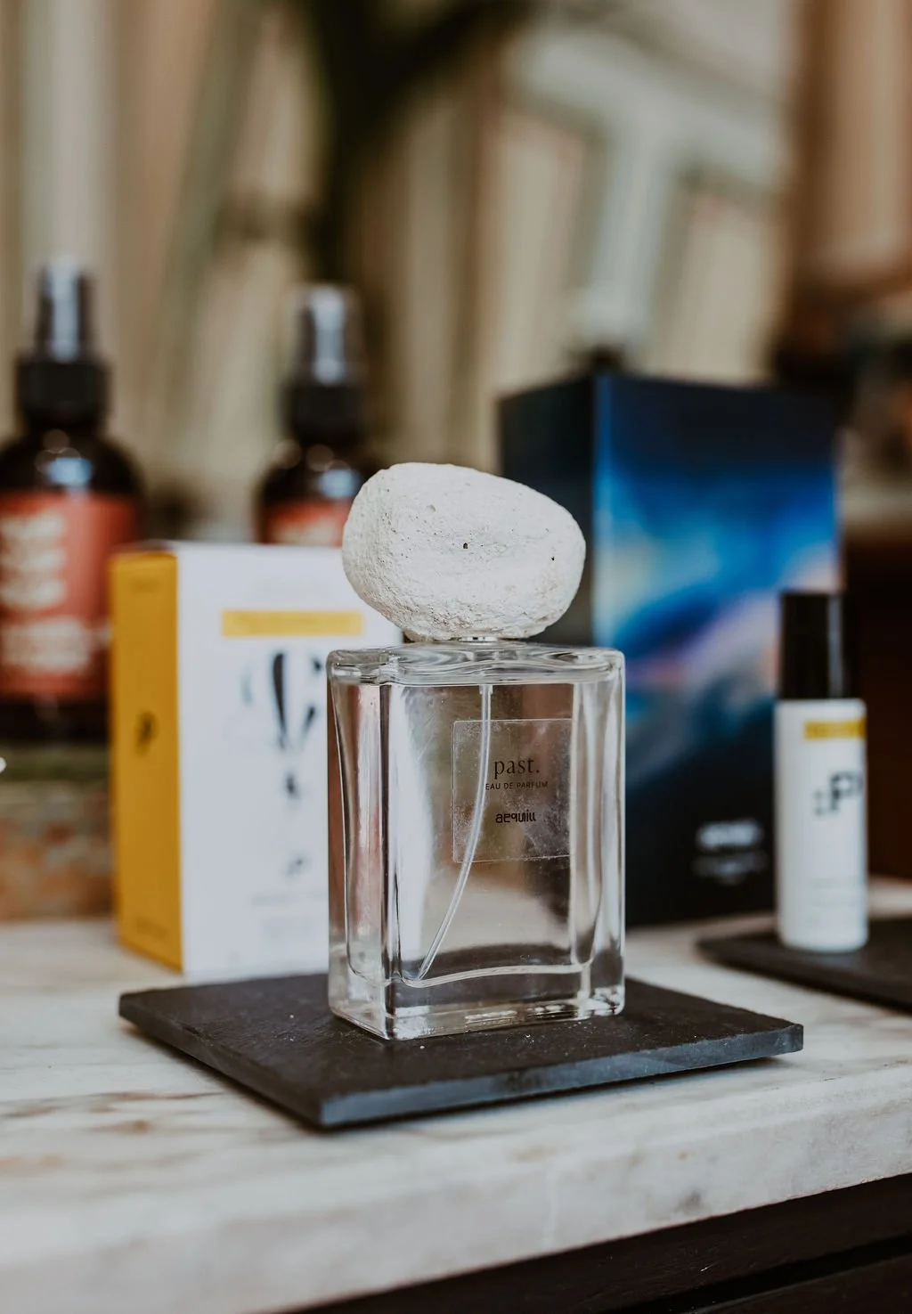 A clear glass perfume bottle with a white stone on top, placed on a black square in front of various cosmetic bottles and boxes.