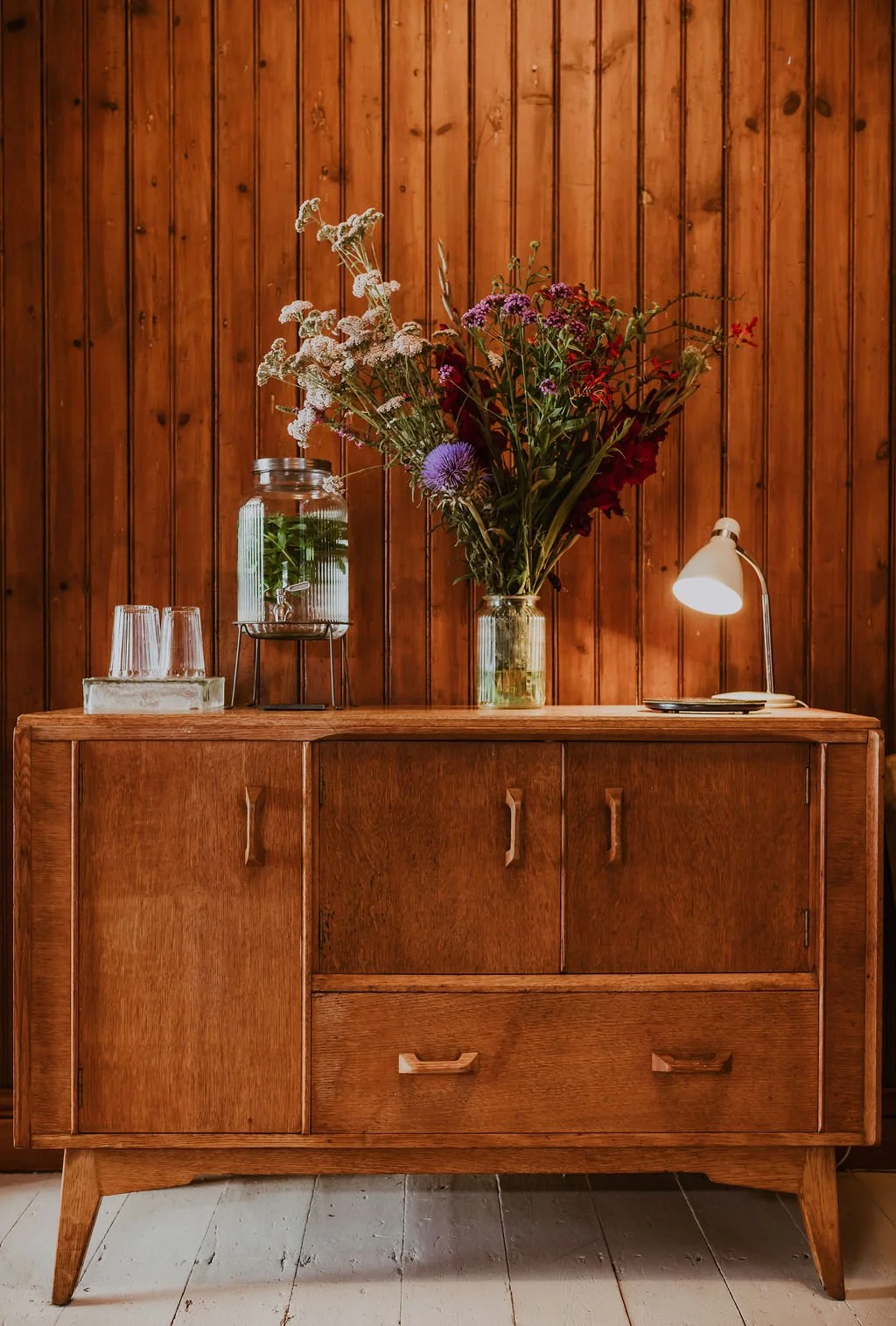 A wooden sideboard against a wood-paneled wall holding a bouquet of mixed flowers in a glass vase, a glass water jar with a metal stand, three glasses, and a white desk lamp.