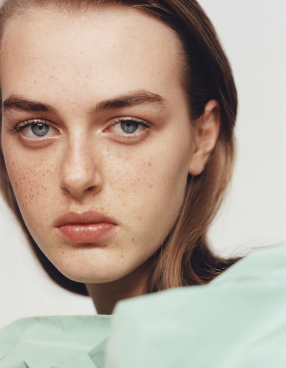 Close-up of a young woman with fair skin, light freckles, blue eyes, and auburn hair, wearing a light green garment.