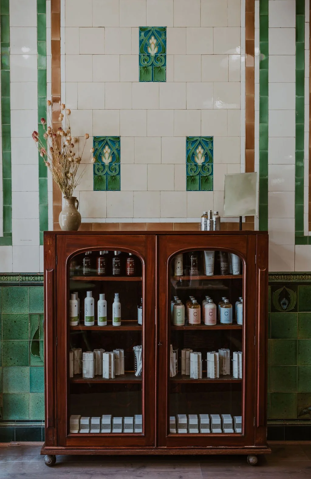 A wooden display cabinet filled with skincare or cosmetic products, topped with a mirror and metallic containers, standing against a decorative tiled wall with green and white patterns and stained glass accents, with a vase of dried flowers nearby.