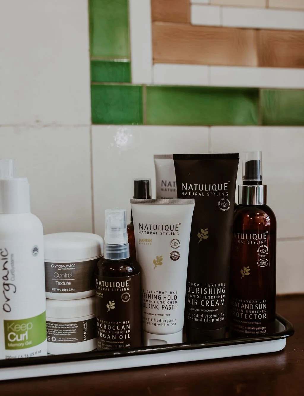 A collection of hair and skincare products on a black tray, including bottles, jars, and tubes, with a decorative tile and wooden shelf in the background.