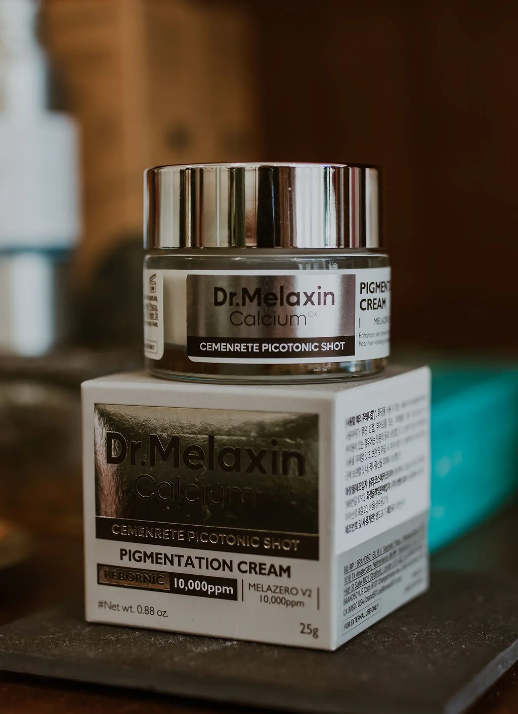 A jar of Dr. Melaxin Calcium pigment pigmentation cream atop a white box, both on a wooden surface, with some blurred objects and a teal item in the background.
