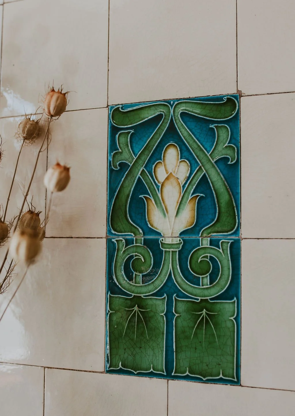 Decorative ceramic tiles with a floral and scroll pattern in green, yellow, and blue, on a tiled wall, with dried flowers nearby.