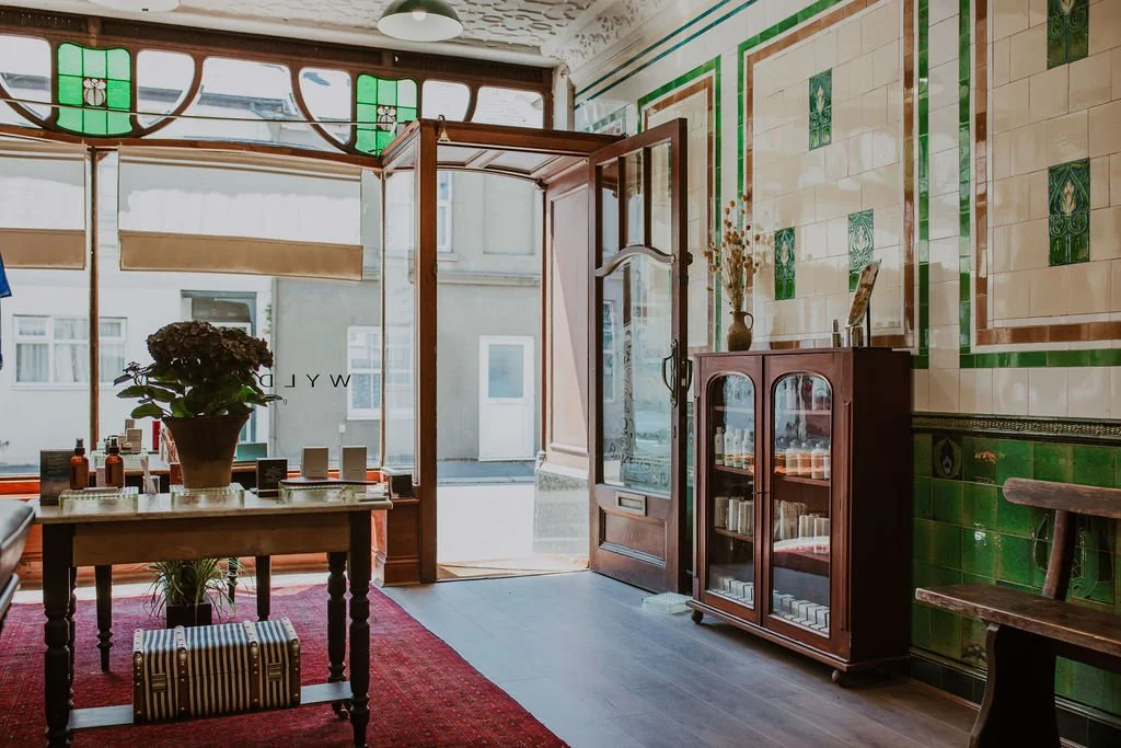 Interior of a vintage-style shop with wooden furniture, a glass display cabinet, a wooden chair, and a table with a potted plant, green and cream tiled walls, and an open glass door revealing a street outside.