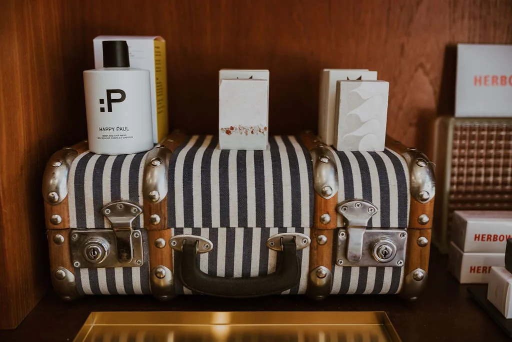 A vintage striped suitcase with metal clasps on a wooden surface, holding a white bottle labeled 'Happy Paul' and some small boxes.