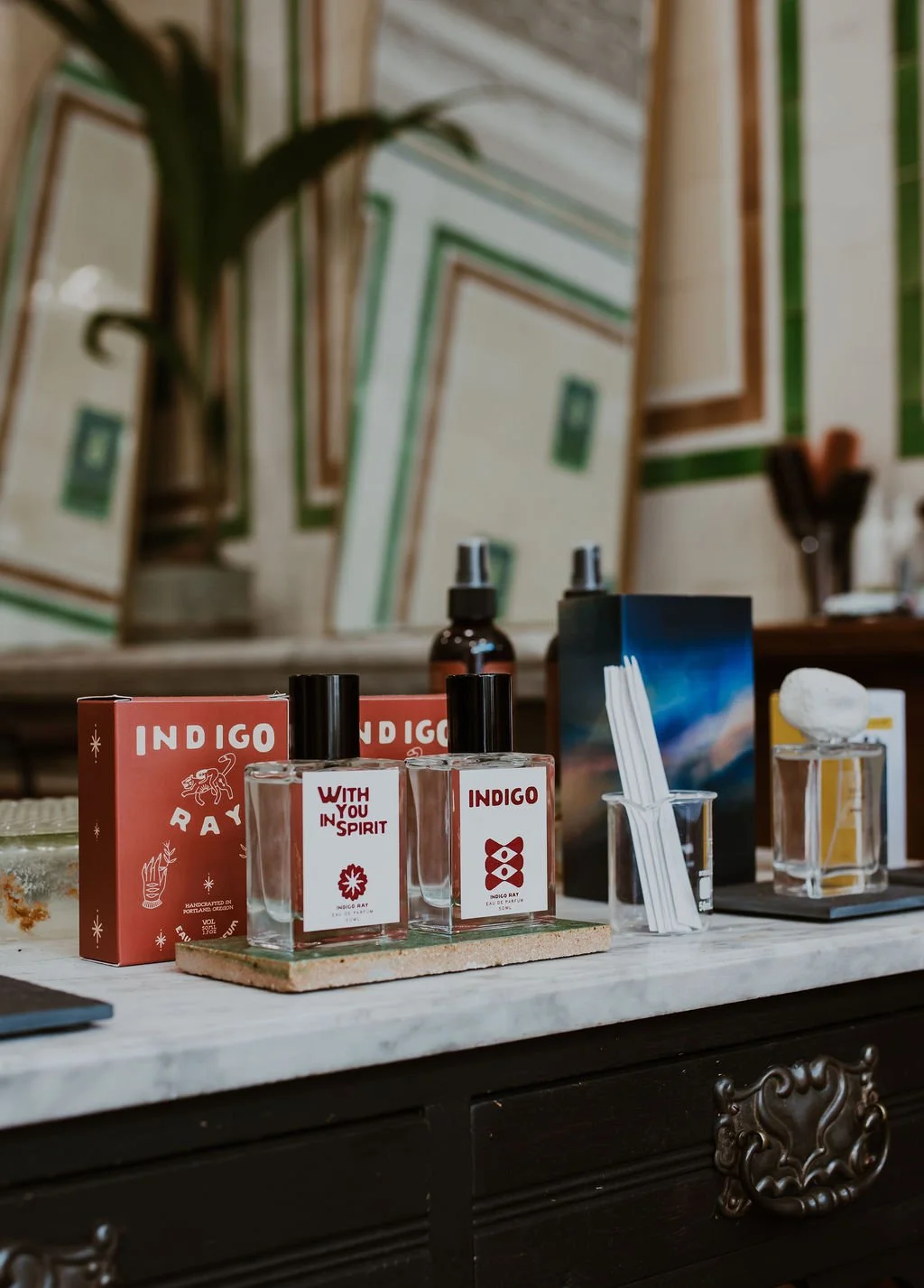Display of skincare or fragrance products on a table, including bottles labeled 'Indigo' and packaging, with artwork and plants in the background.