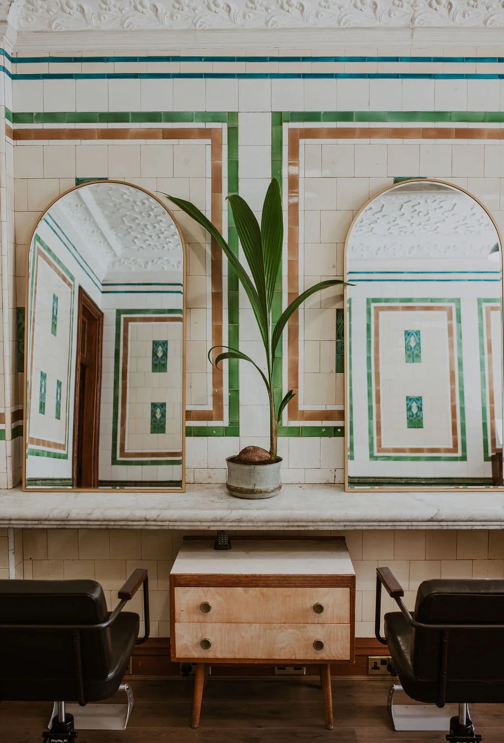 Interior with two large arched mirrors, a potted plant between them, and a small wooden dresser underneath the mirror. The wall behind has decorative tile patterns in green, brown, and cream. Two black chairs are in front of the dresser.
