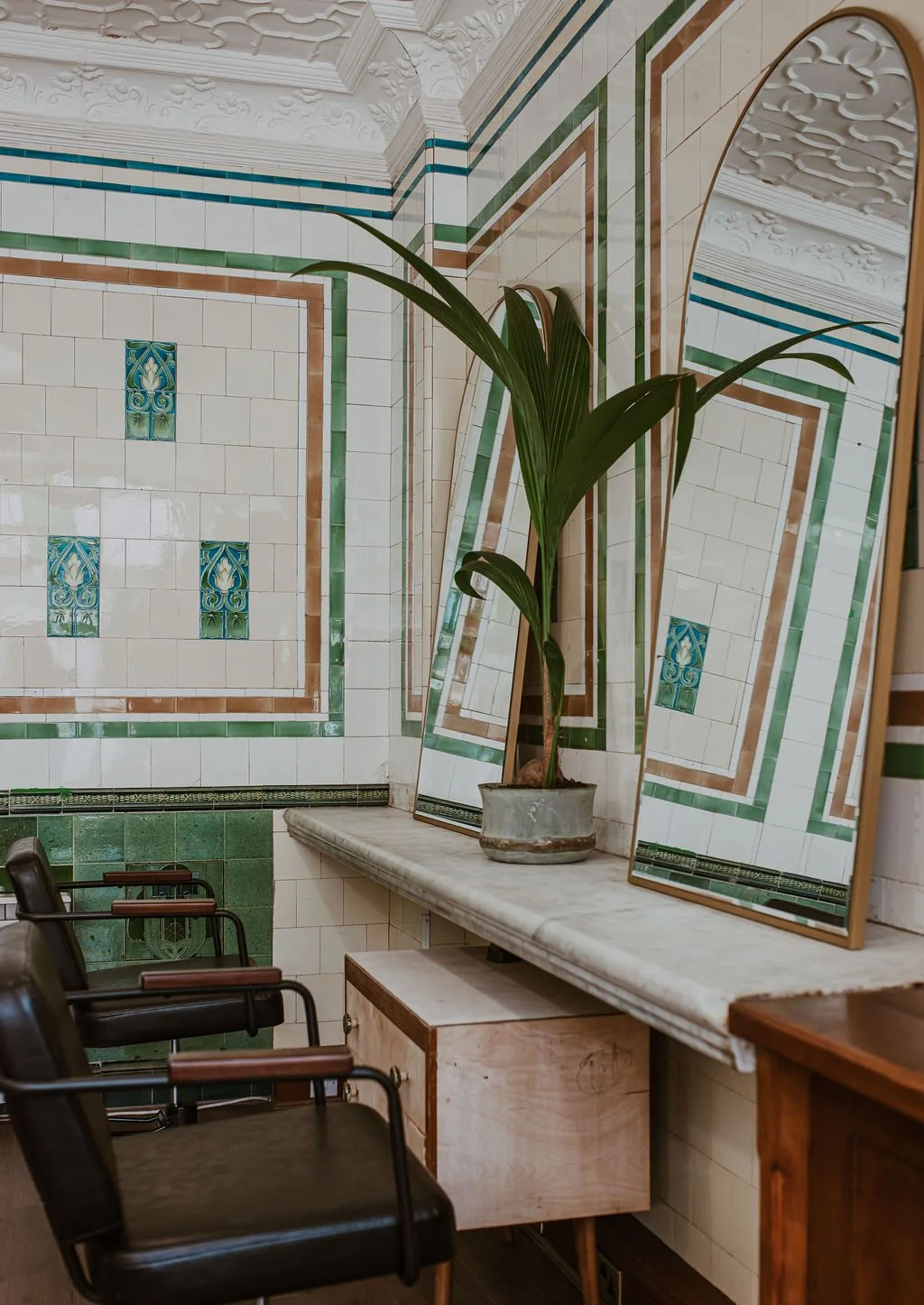 Interior of a room with tiled walls, a marble-topped table, a potted plant in front of three mirrors, and a couple of chairs with wooden armrests.