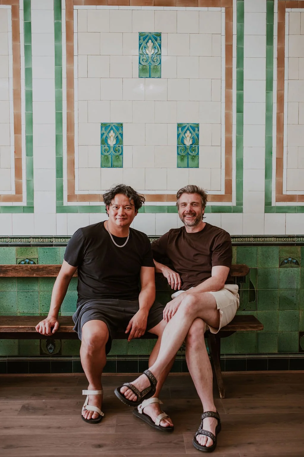 Two men sitting on a wooden bench in front of a decorative tiled wall with green and cream tiles, smiling and relaxing.