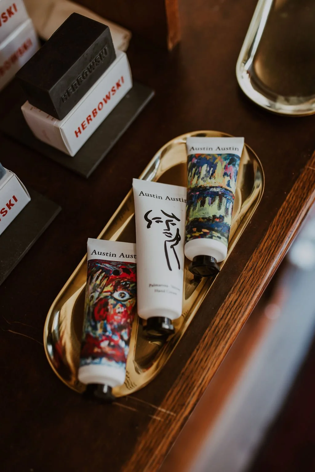 Three tubes of Austin Austin hand cream with artist-designed packaging on a gold tray, near HERBORAGE soap bars and a mirrored object on a dark wooden surface.