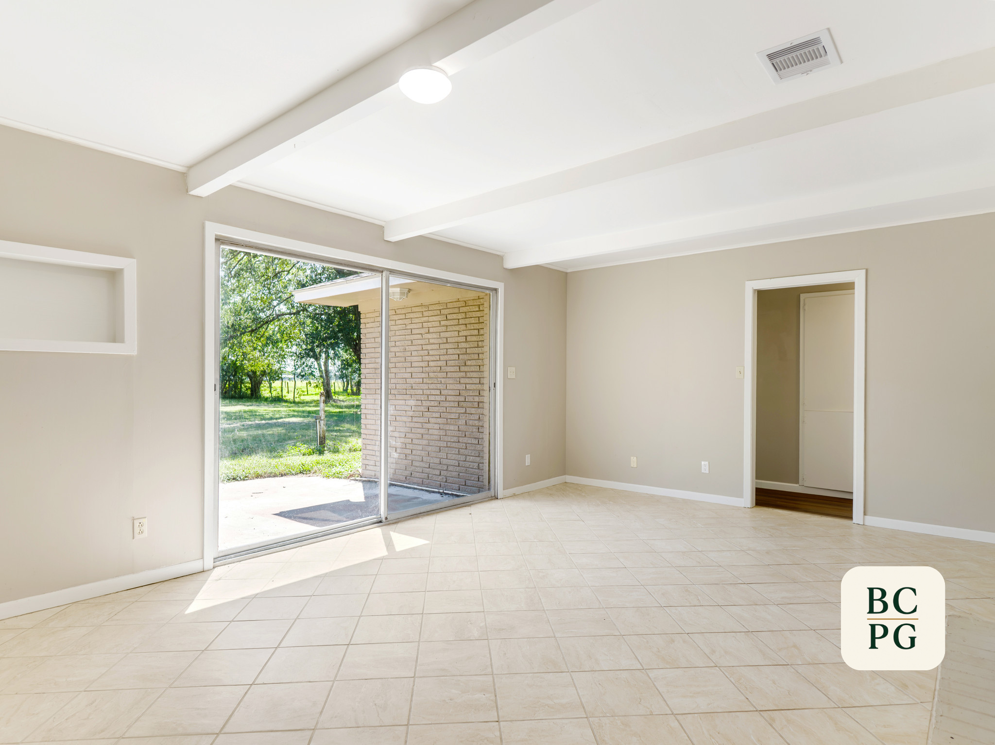 Empty room with beige tiled floor, light-colored walls, large sliding glass door leading outside, and two doorways. Sunlight streams into the room, revealing a view of green trees and grass outside.