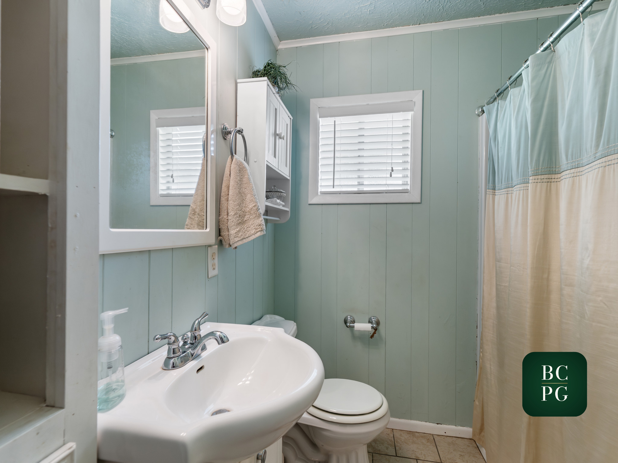 A small bathroom with light green paneled walls, two windows with white shutters, a white sink with a silver faucet and soap dispenser, a beige towel hanging on a silver round towel ring, a white cabinet above the toilet with a plant on top, a toilet