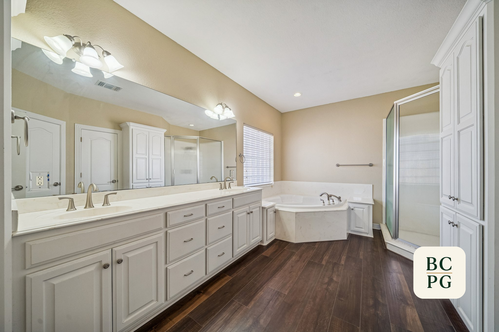 A spacious modern bathroom featuring a double sink vanity with white cabinetry, a large mirror, and multiple light fixtures. There is a corner bathtub and a separate glass-enclosed shower unit. The bathroom has a window with blinds, and the floor is 