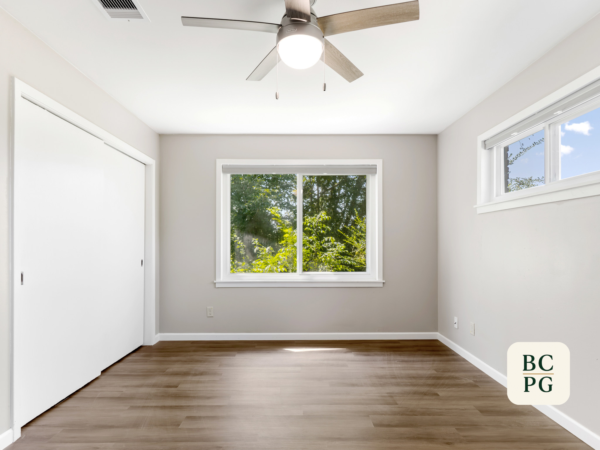 Empty room with white walls, hardwood floor, two large windows, and a ceiling fan with light.