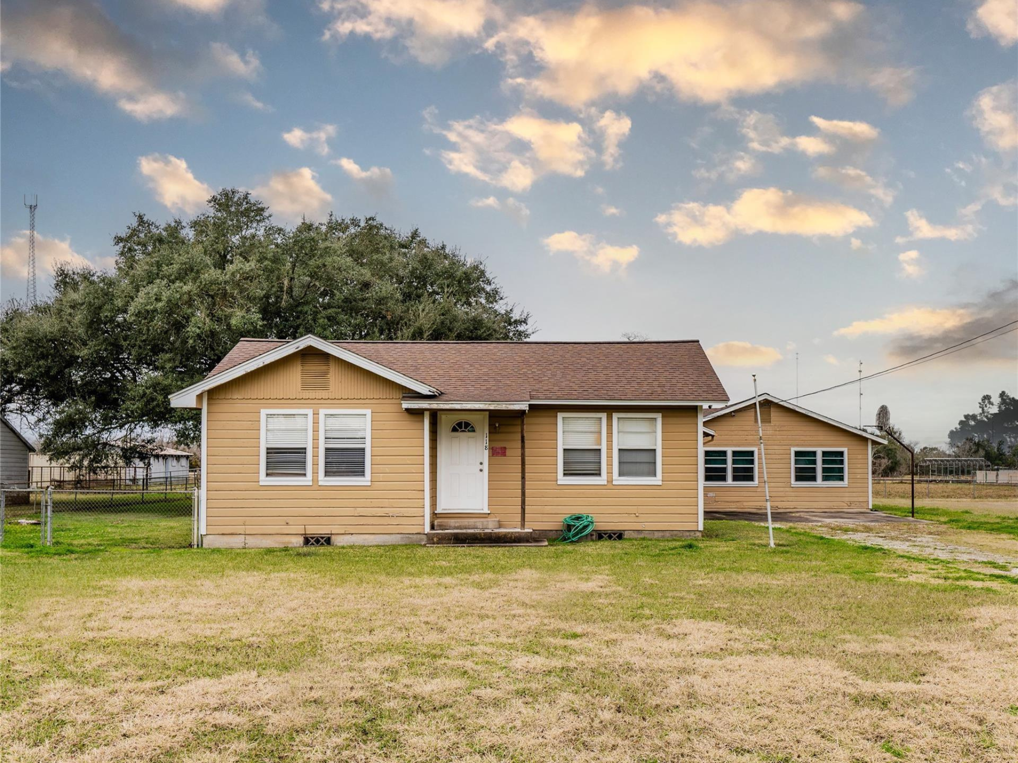 A yellow house with white trim and a brown shingle roof, two windows, and a gray front door, with a hose reel nearby and a large tree in the background. The sky is partly cloudy with light clouds and blue sky. There is a grassy yard and neighboring houses visible.