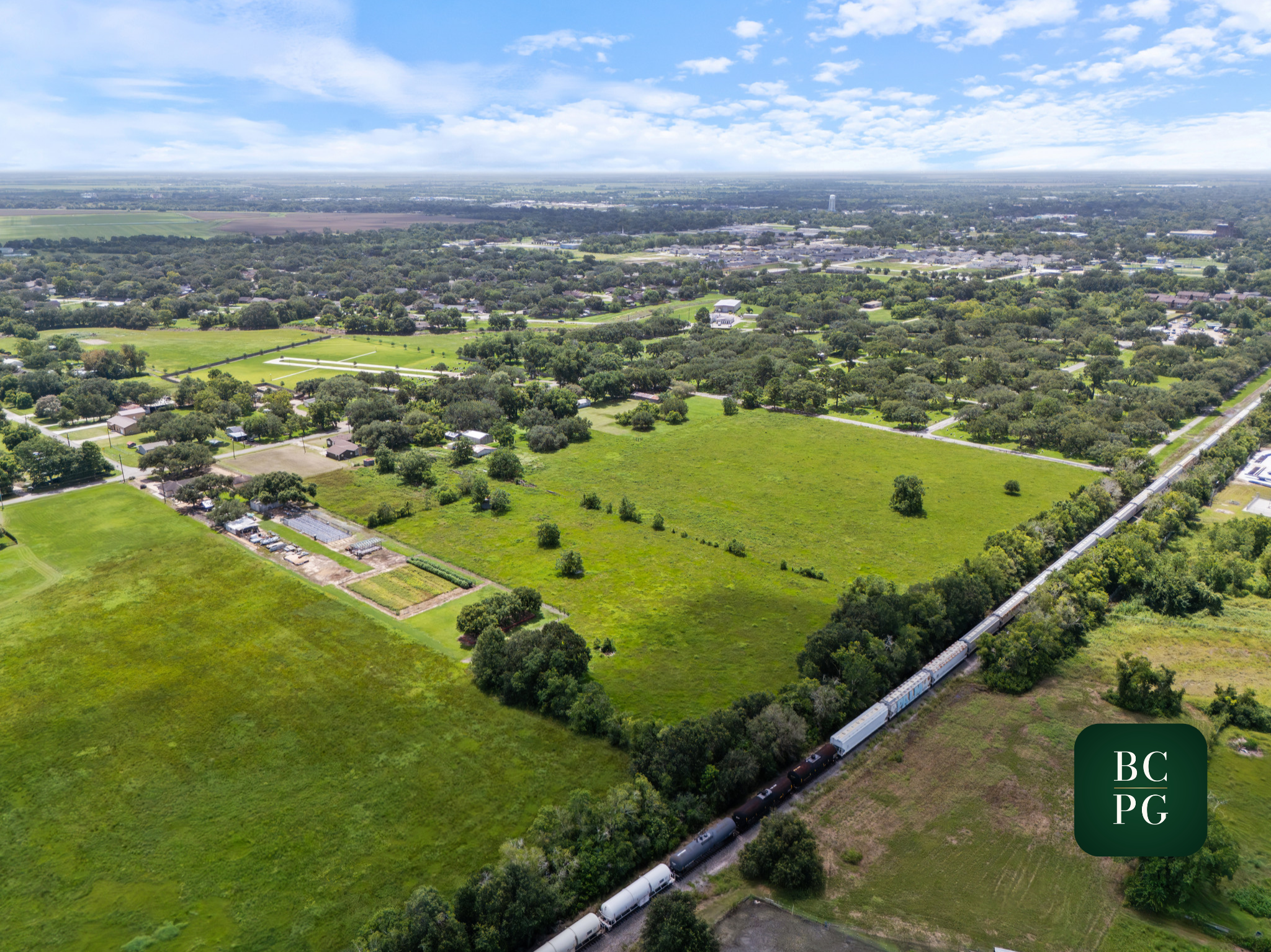 An aerial view of a green suburban area with houses, trees, and open fields, with a train passing through on the right side under a partly cloudy sky.