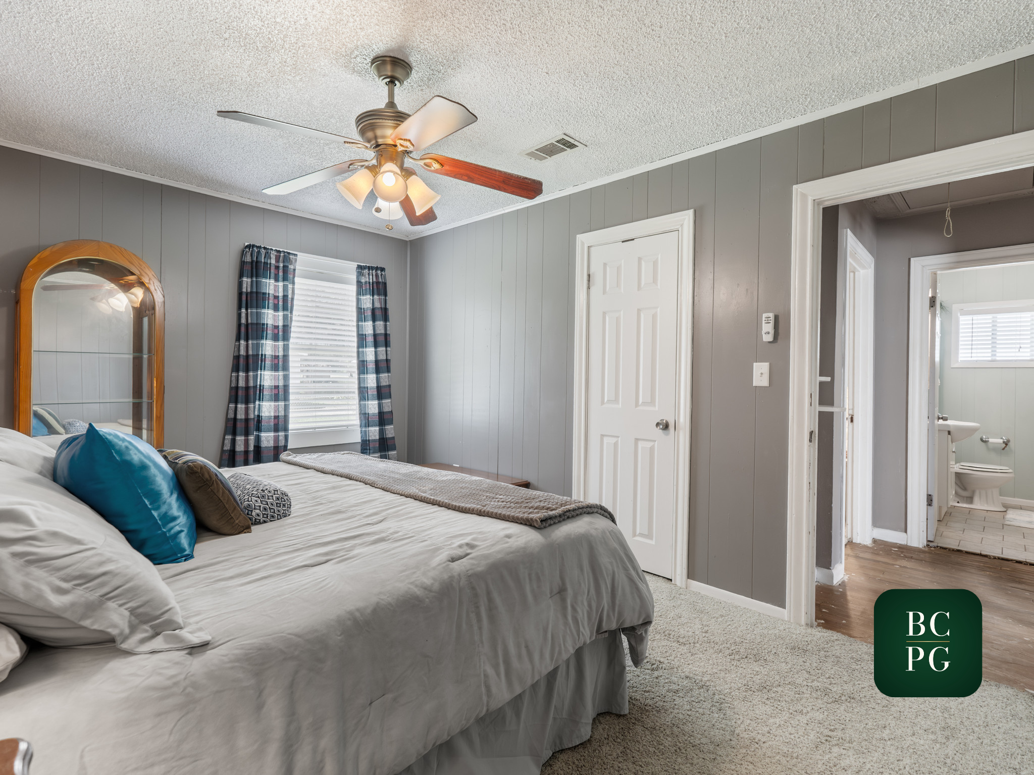 A bedroom with a white bed and pillows, gray walls, a window with plaid curtains, and a ceiling fan with lights.