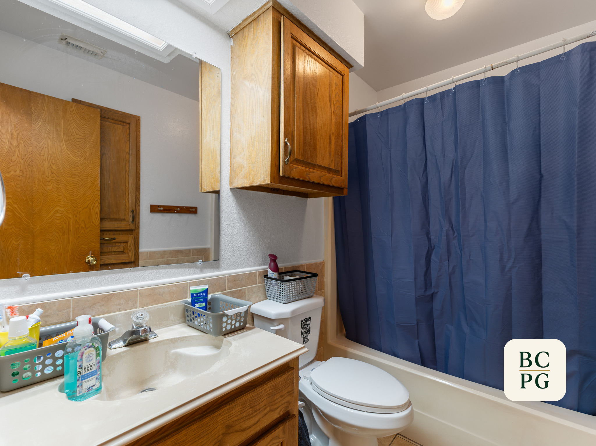 A bathroom with a wooden cabinet above the sink, a mirror, and a blue shower curtain. The sink has various toiletries including hand soap and toothbrushes, and there is a toilet next to the shower. The bathroom has beige tile flooring and white walls