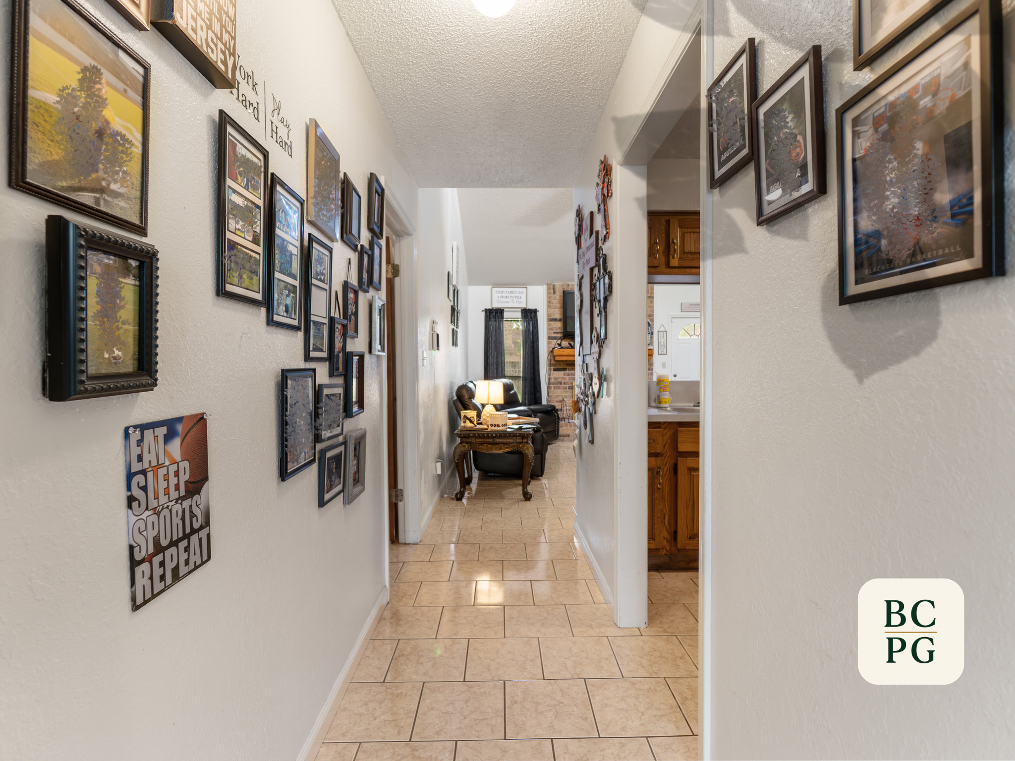 A hallway with tiled floor and white textured walls. Multiple framed pictures and posters decorate the walls, including a motivational quote. There is a view through a doorway into a room with a brick wall, a window with black curtains, and a small t