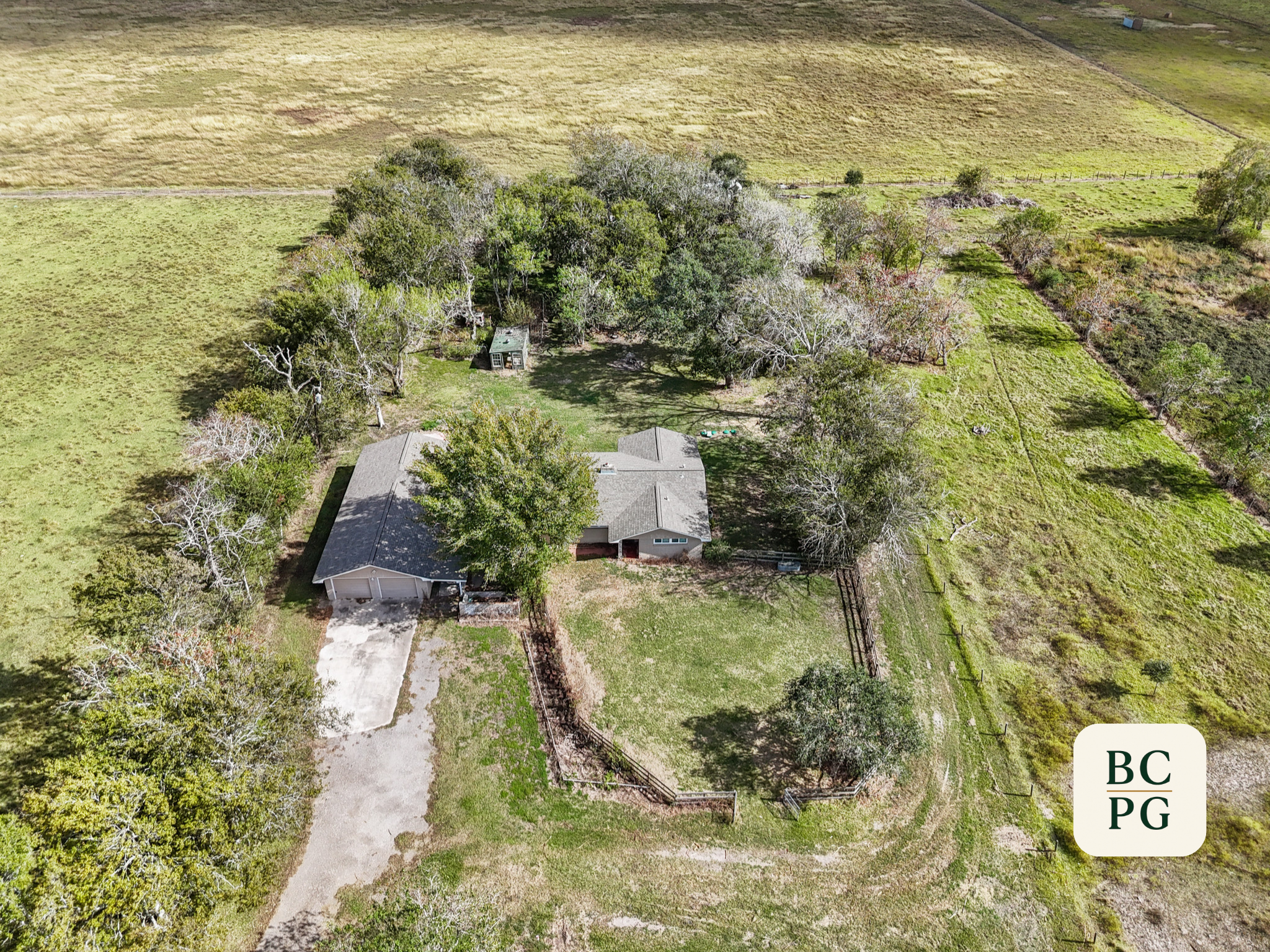 Aerial view of a suburban backyard with trees, a house, a driveway, and expansive grassy fields in the background.
