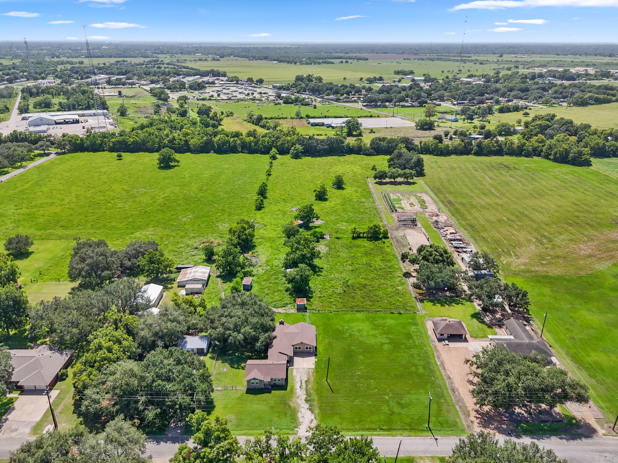 Aerial view of a rural area with green fields, trees, houses, and farm structures under a partly cloudy sky.