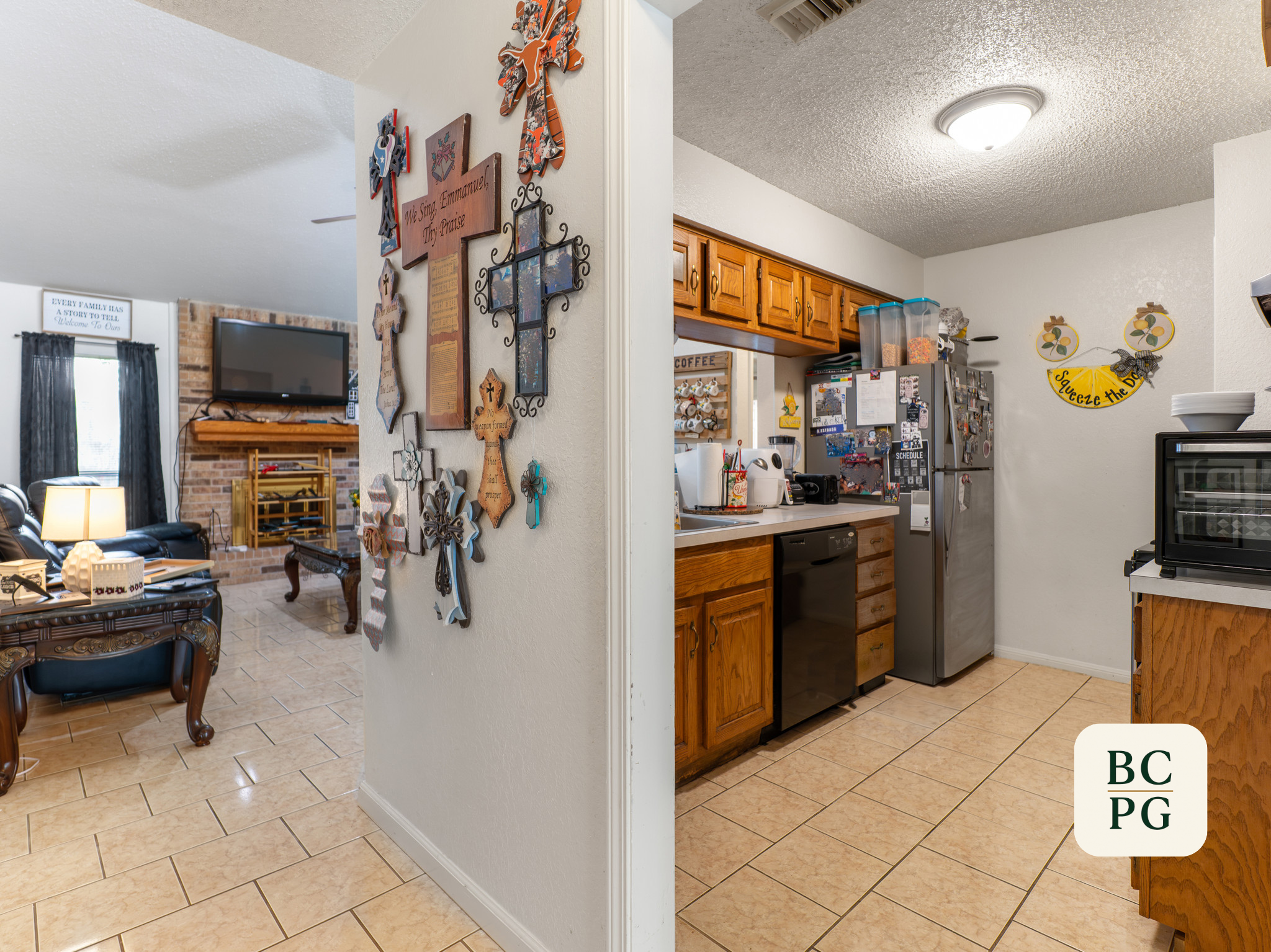 Interior view of a kitchen and living room separated by a wall with various wall decorations. The kitchen has wooden cabinets, a refrigerator, and a countertop with containers and appliances. The living room features a TV mounted on a brick fireplace