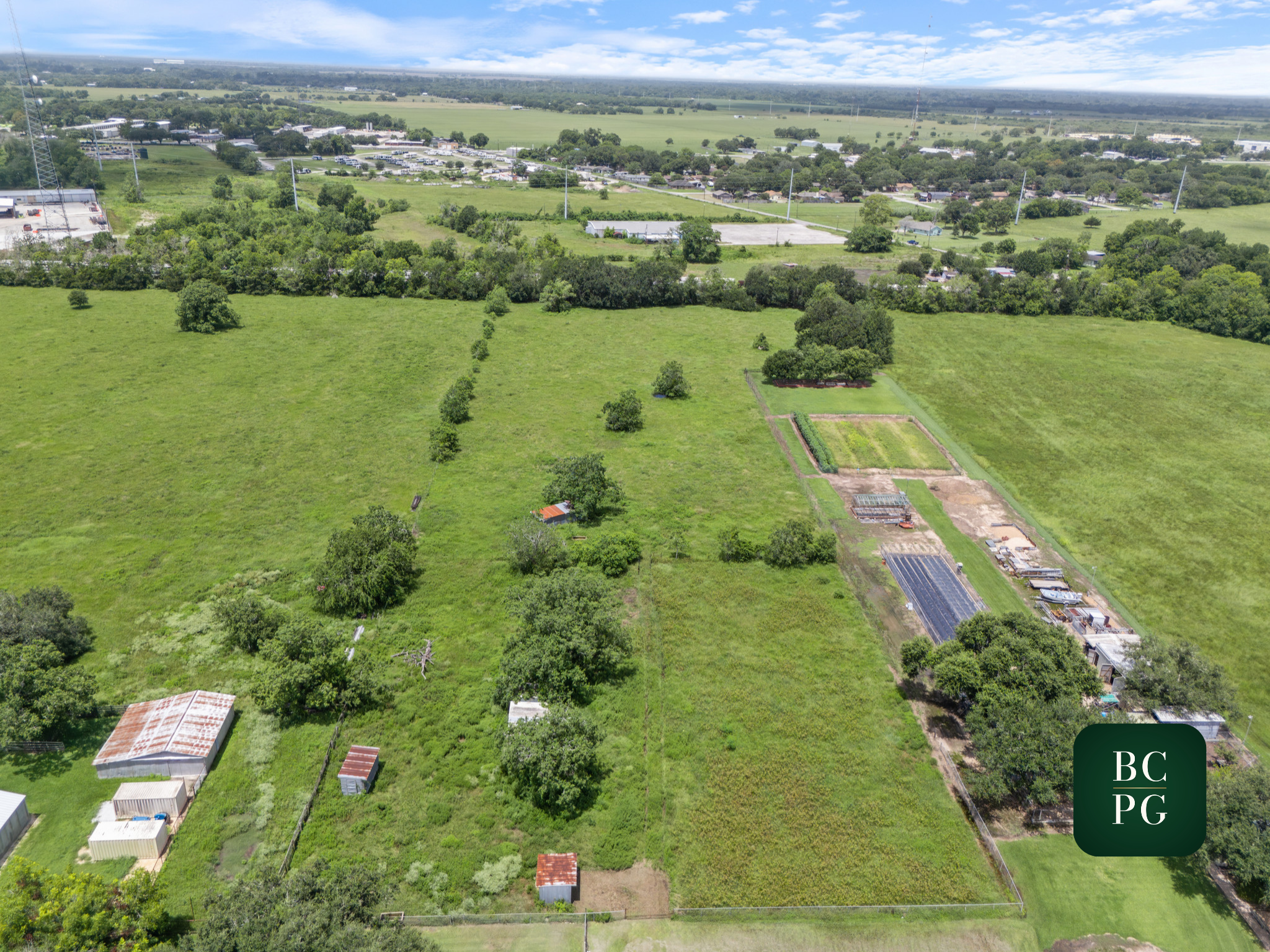 Aerial view of a green landscape with open fields, trees, small buildings, and a distant urban area with parking lots and roads under a partly cloudy sky.